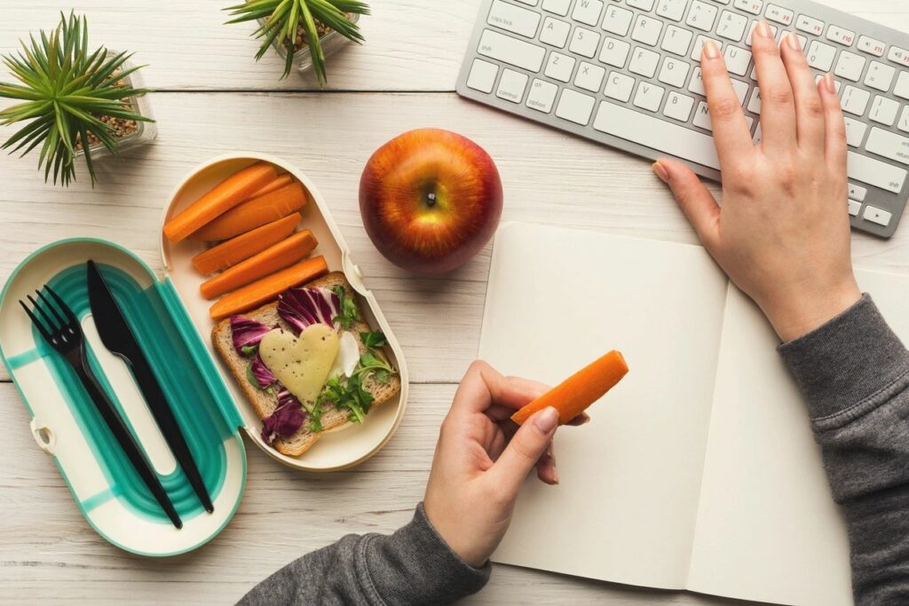 Healthy lunch with apples, carrots, and sandwich at the workplace with employee using keyboard and eating.