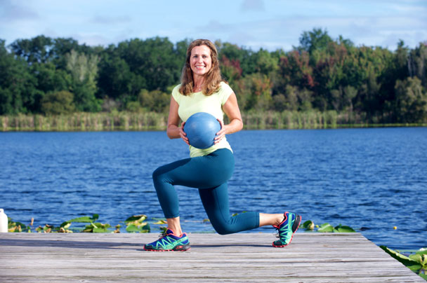 Debbie Ivie holding a medicine ball in a twisted lunge position on a dock by the lake.