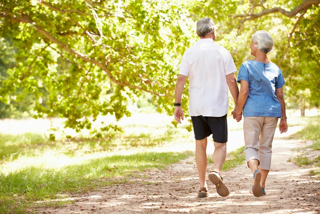 Functionally aging man and woman taking a walk while holding hands on a dirt path with trees around.