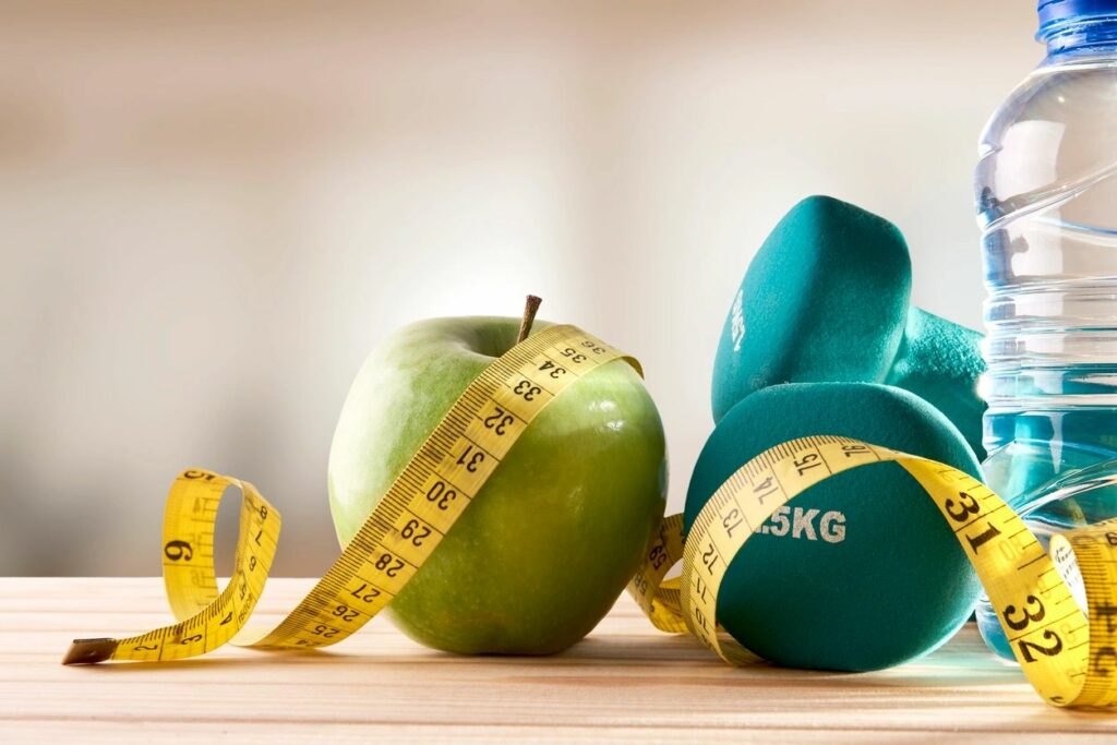 Healthy photo with green apple, turquoise weights, bottle of water, and yellow measuring tape.